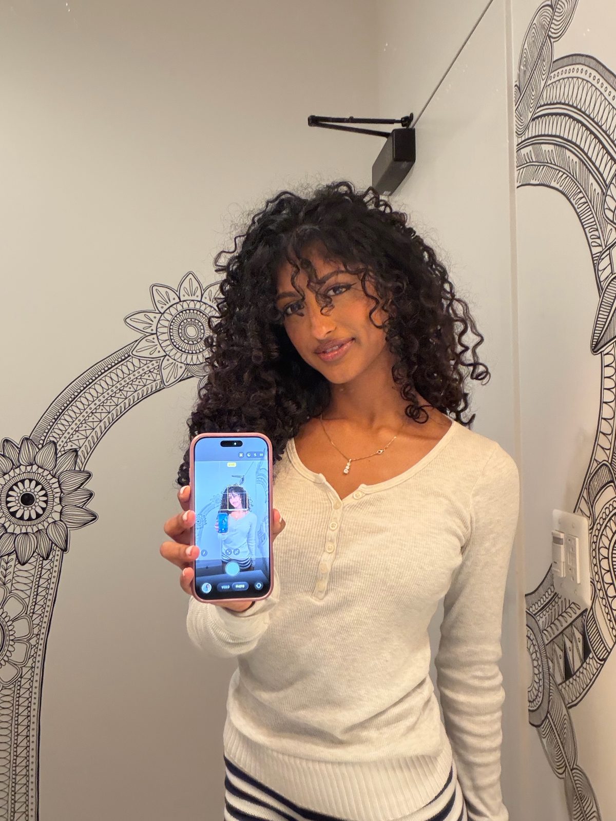 Mirror selfie of Jerusha in a cream henley, her curls up close and defined