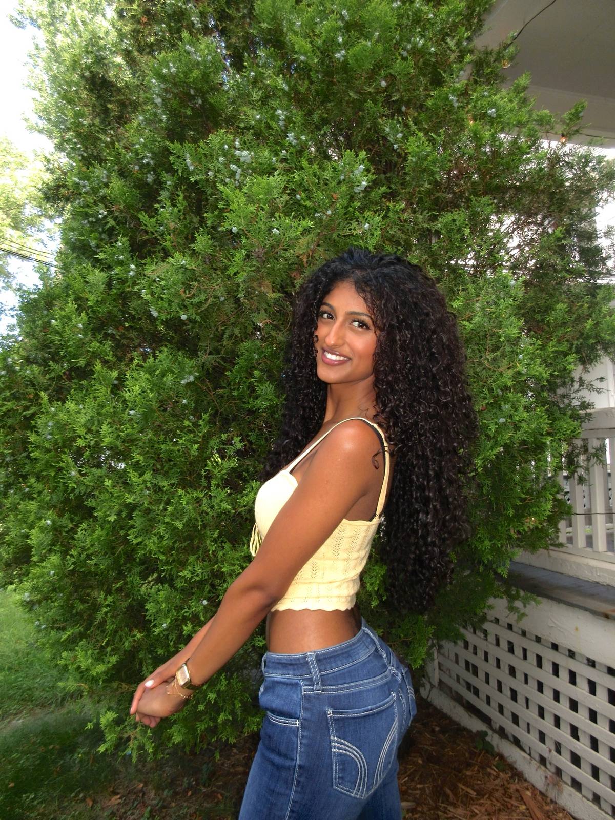 Jerusha outdoors by green hedges in a yellow top and jeans, long curls down her back